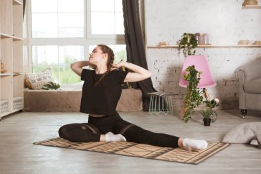 Evdeki odada esneyen ve yoga egzersizi yapan genç bir kadın. Evde fitness, aktif giyim. Sağlıklı ve spor yaşam tarzı. 