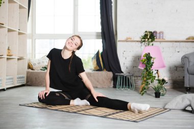 Evdeki odada esneyen ve yoga egzersizi yapan genç bir kadın. Evde fitness, aktif giyim. Sağlıklı ve spor yaşam tarzı. 