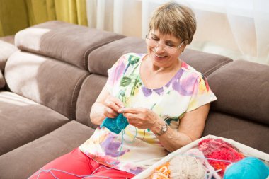 Yaşlı bir kadın odada bir kanepede oturmuş örgü örüyor.