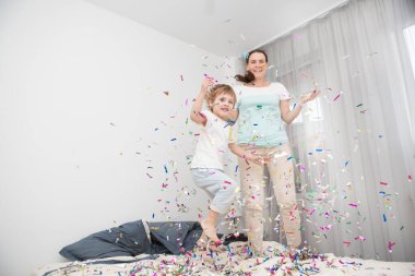  Anne ve oğlu konfeti patlatarak kutluyorlar. Olumlu duygular. Mutlu küçük çocuk ve annesi yatakta zıplıyorlar.