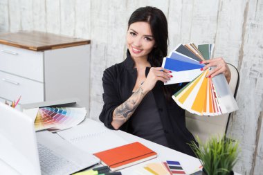 Successful business woman designer with a tattoo  sitting at the desk with instruments, colour samples and laptop in office