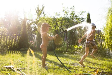 Mutlu çocuklar erkek ve kız kardeşler bahçe hortumuyla oynuyorlar ve güneşli arka bahçede su püskürterek eğleniyorlar. Yaz zamanı..