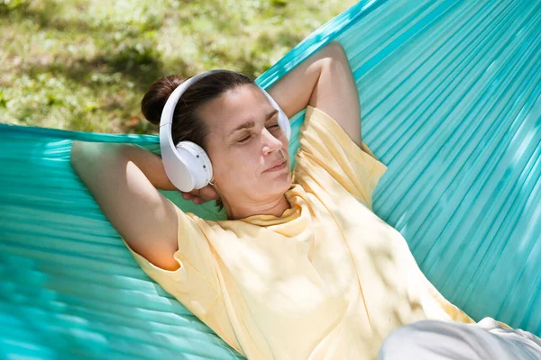音频治疗 戴耳机的女人在夏季公园或花园的吊床上听电话录音 图库图片