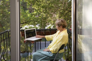 Kıdemli kadın bir yaz günü evinin balkonunda dizüstü bilgisayar kullanıyor. yaşlı yaşam tarzı.