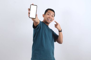 Heyecanlı genç Asyalı adam elinde tutuyor ve beyaz ekranlı bir cep telefonunu işaret ediyor. Uygulama ya da web sitesi reklamı için mükemmel. Beyazda izole edilmiş.