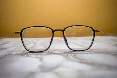 Açık mermer yüzey ve bej zemin üzerine yerleştirilmiş ince tozlu siyah kare gözlük, günlük gerçekçilikle minimalist moda ve görüş netliğini birleştiriyor.