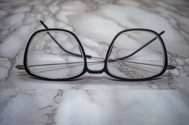 Mermer bir yüzeyde duran şeffaf mercekli klasik siyah kare gözlükler, minimalist kompozisyonda zamansız zarafet, berraklık ve klasik moda cazibesini sergiliyor.