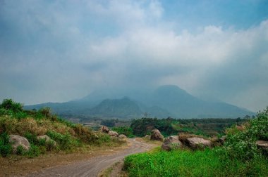 Penanggungan Dağı, yeşil bitki örtüsü, büyük kayalar ve bulutlu bir gökyüzünün altındaki dolambaçlı toprak yolu ile Endonezya 'nın verimli bir arazisi boyunca uzanır. Doğa, seyahat ve manzara teması için mükemmel.
