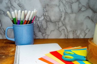 Mavi bir kupa, defter, makas ve kağıt renkli işaretleri mermer bir arka plan karşı ahşap bir masa üzerinde, sanat malzemeleri ve yaratıcı araçlar sergilenen.