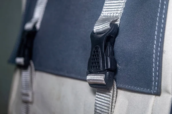 Siyah plastik tokalı ve gri kayışlı gri beyaz bir sırt çantasının yakın çekimi. Sırt çantası görünür dikişli ve desenli yüzeyi olan kumaştan yapılmıştır..