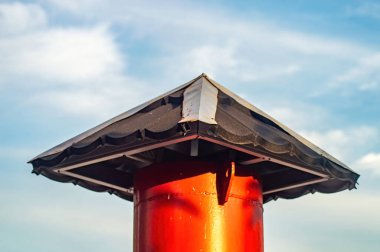 Kare çatısı olan kırmızı metal bir direk, yumuşak bulutlu mavi bir gökyüzüne karşı duruyor. Açık havada basit ama çarpıcı bir mimari detay oluşturuyor..