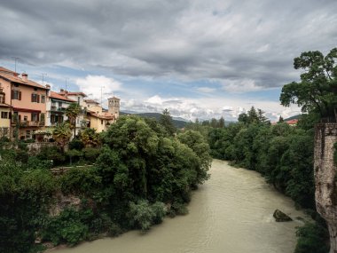 View of the River Natisone winding through lush greenery and colorful houses in Cividale del Friuli, Italy. Overcast skies create a dramatic atmosphere above the peaceful riverside town.