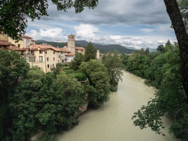 View of the River Natisone winding through lush greenery and colorful houses in Cividale del Friuli, Italy. Overcast skies create a dramatic atmosphere above the peaceful riverside town.