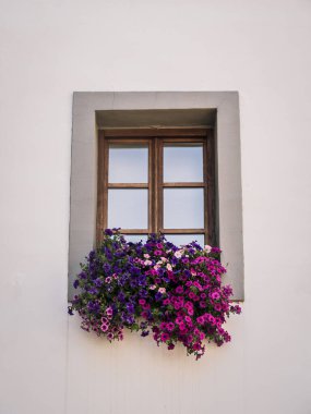 A rustic wooden window framed by a simple white wall, decorated with a vibrant arrangement of purple and pink flowers. The scene evokes charm and elegance in a minimal architectural style.