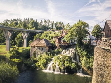 Hırvatistan 'ın Rastoke kentini taş su değirmenleri, teraslı şelaleler, yemyeşil yemyeşil alanlar ve manzaralı bir çerçevede yakın modern bir köprü ile geniş bir manzara.
