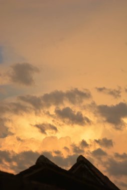 Bir evin çatısının koyu gölgesini, yumuşak kümülüs bulutlarıyla dolu, çarpıcı, turuncu gün batımı gökyüzüne karşı gösteren dikey bir çekim.