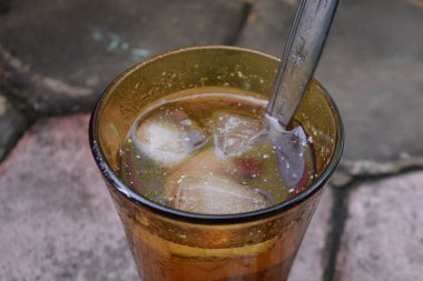 Soğuk bir bardak buzlu çaya yakın, görünen buz küpleri ve metal bir kaşıkla dışarıda taş bir kaldırımda fotoğraflanmış. Doğal ışık, yoğunlaşma ve otantik sokak yemekleri onu ferahlatıcı, yaz sıcağı ve Endonesi için ideal kılıyor.