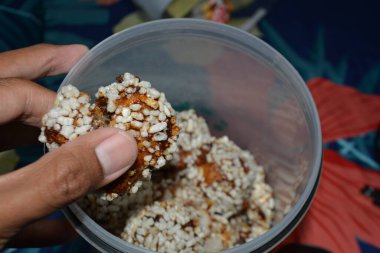 Bir el, benzer şekerlerle dolu bir konteynırın üzerinde tek bir şişirilmiş pirinç topu sunar. Çıtır tanelerin ve şurup dokusunun net ayrıntıları servis ve seçim temalarını destekler.