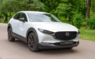 Park yolunda beyaz Mazda CX-30 marka kompakt bir SUV. Çerçevesi yemyeşil ağaçlarla çevrili. Pürüzsüz ızgara ve modern tasarım, kentin çok yönlülüğünü ve açıkhava hazırlığını sakin ve doğal bir ortamda iletiyor. Ukrayna, Kiev 05 / 29 / 2024