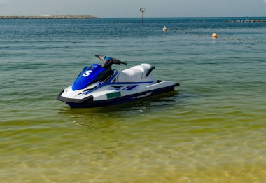 Mavi ve beyaz bir jet ski kumlu bir sahilde sakin bir suya park edilmiştir. Güneş batıyor ve temiz bir su manzarası yaratıyor. Yakınlarda bazı şamandıralar görülüyor..