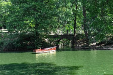 Yeşil bir gölde ahşap bir tekne. Arka planda yemyeşil ağaçlar ve bir taş köprü görülebilir. Parlak güneş ışığı çevreyi aydınlatır ve doğal çevreyi açıkça gösterir..
