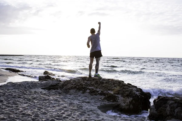 Deportivo hombre de pie en la roca con levantado en el aire puño amanecer en la playa — Foto de Stock