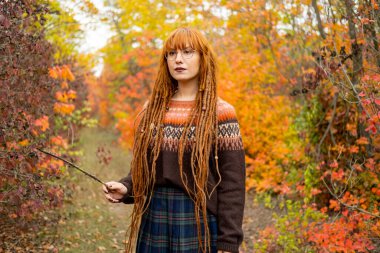 Kızıl saçlı genç bir kadın ve güzel sonbahar ormanlarında süveter giyiyor.