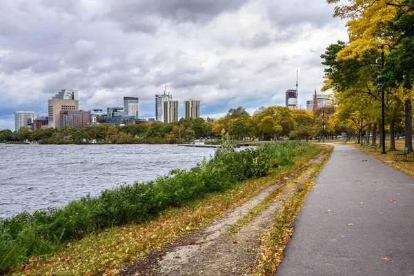 Empty riverside path lined with ebches and autumn trees in a public park under storm clouds. High rise downtown buildings are visible in background. Boston, MA, USA.