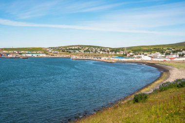 Açık bir yaz gününde limana bakan yamaç kenarındaki evleri olan güzel küçük bir sahil kasabası. Ön planda terk edilmiş bir plaj görülebilir. Husavik, İzlanda.