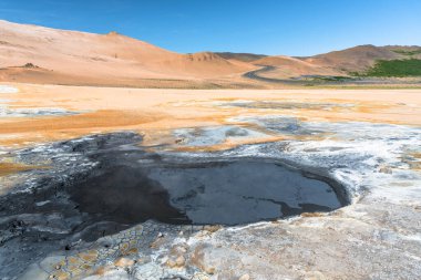 Açık bir yaz gününde İzlanda 'nın bir jeotermal bölgesinde kaynayan çamur havuzunun fotoğrafı. Arka planda çorak bir volkanik tepeye tırmanan bir yol görülebilir. Hver, İzlanda.