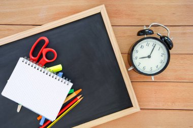 Tahta bir masanın üzerindeki küçük bir tahtada çalar saatle birlikte açık defter ve renkli okul malzemeleri. Uzayı kopyala Okul konseptine dönelim..