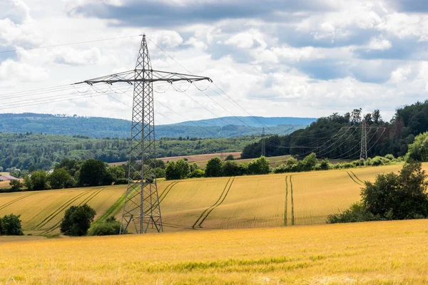 曇りの夏の日に栽培分野の電気パイロン 森の丘が背景にあります ドイツの田舎 — ストック写真