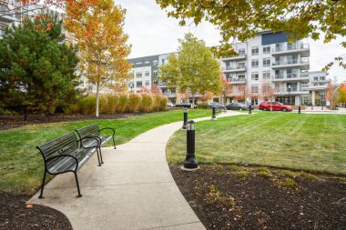Bulutlu bir sonbahar sabahında modern apartman bloklarıyla çevrili bir parkta asfalt yol boyunca uzanan boş metal banklar. Boston, MA, ABD.