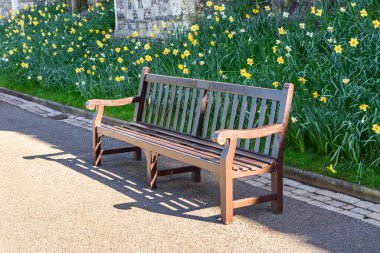 Parkın asfalt yolu boyunca boş ahşap bir bank. Arka planda bir çiçek yatağı var. Windsor, İngiltere, İngiltere.