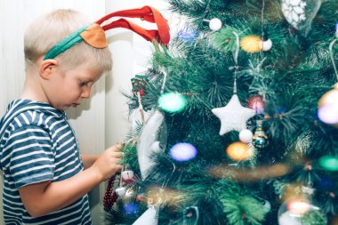 Kafasında geyik boynuzları olan sevimli küçük çocuk Noel ağacını süslüyor.