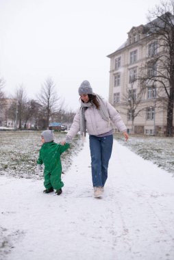 Genç anne, kış parkında karlı bir yolda yürürken bebeğiyle el ele tutuşuyor. Sıcak aile anları, doğal yaşam tarzı, ebeveynlik ve çocukluk konsepti..