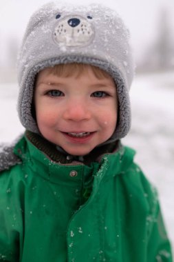 Karlı bir günde kışın giyinmiş gülümseyen bir çocuğun yakın plan portresi. Doğal ışık, yumuşak arka plan, rahat kış çocukluğu konsepti.