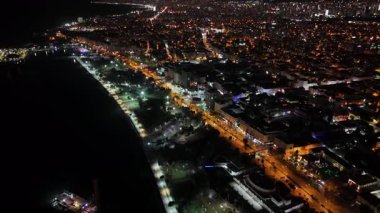 Hava aracı gece vakti aydınlık bir sahil kenti boyunca uçarken karanlık denizin yanındaki binaların ve sokakların aydınlık şehir ışıklarını ve kıyı şeridini gösteriyor.