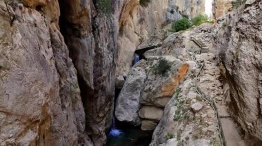 Dar bir kanyonun nefes kesen güzelliği, yükselen kaya duvarlarına oyulmuş çağlayan bir şelale ve engebeli bir patikayı gösteren havadan bir bakış açısıyla ortaya çıkar.