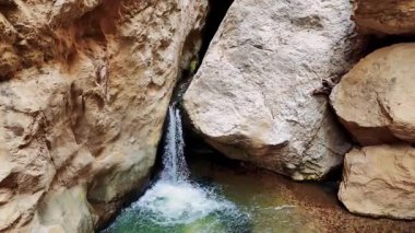 Dar kayalık bir geçitten sızan sakin bir şelale. Kristal berrak su, pürüzsüz taşların üzerine nazikçe yuvarlanarak ışıldayan bir turkuaz deposuna dönüşür.