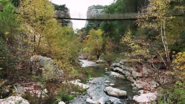 Göz kamaştırıcı bir hava perspektifi dar bir dağ geçidinde geriye doğru süzülüyor, aşağıda dönen bir nehir ve yukarıda uzanan bir halat asma köprüsü var.