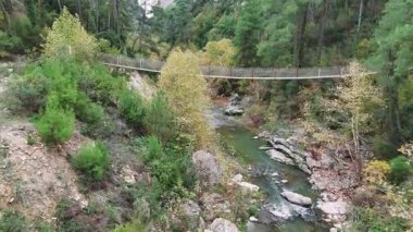 İnsansız hava aracı, çarpıcı bir vahşi doğa ve kristal berrak bir dağ deresi ile çevrili pitoresk bir nehir kanyonu boyunca uzanan taşralı asma halat köprüsünün üzerinde süzülüyor.