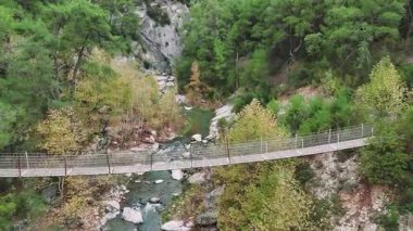 Engebeli bir vadiyi kesen kıvrımlı bir nehrin yer aldığı manzaralı dağ manzarası. Yeşil zümrüt ağaçları, yarığı çevreleyen uzun bir asma köprünün yanında manzarayı işaretliyor.