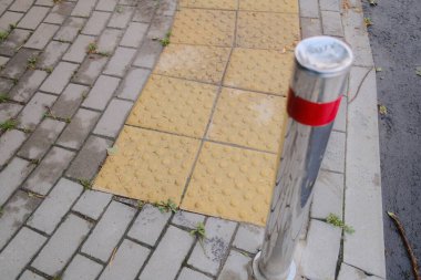 Islak kaldırımda sarı desenli fayanslar ve metal bir kalas var..