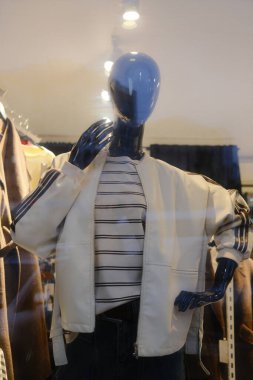 Manken bir giyim mağazasının vitrininde ceket ve gömlek giyer ve el kaldırarak poz verir..