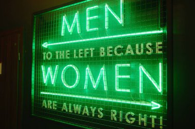 Parlak yeşil neon bir tabela cinsiyete özel tuvalet talimatlarını bir şakayla gösterir.