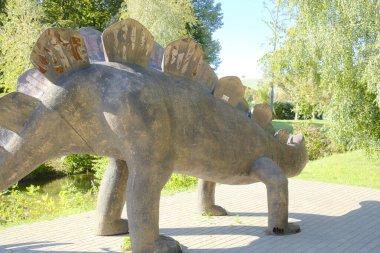 Asfaltlı bir alanda gri bir dinozor duruyor. Arka planda ağaçlar ve yeşil çimenler var..