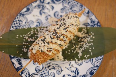 Bol yağda kızartılmış mısırlı sosis, susam tohumlarıyla dekoratif bir tabağa konur..