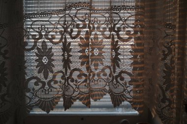 Güneş ışığı ev penceresinden akar, hassas dantel perdelerin ve yatay perdelerin karmaşık desenlerini ortaya çıkarır..