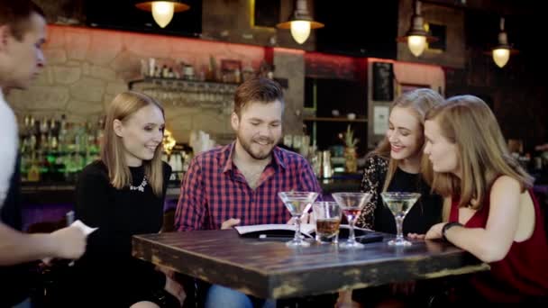 Happy young people sitting in the bar reading menu card and giving orders to the male waiter — Stock Video